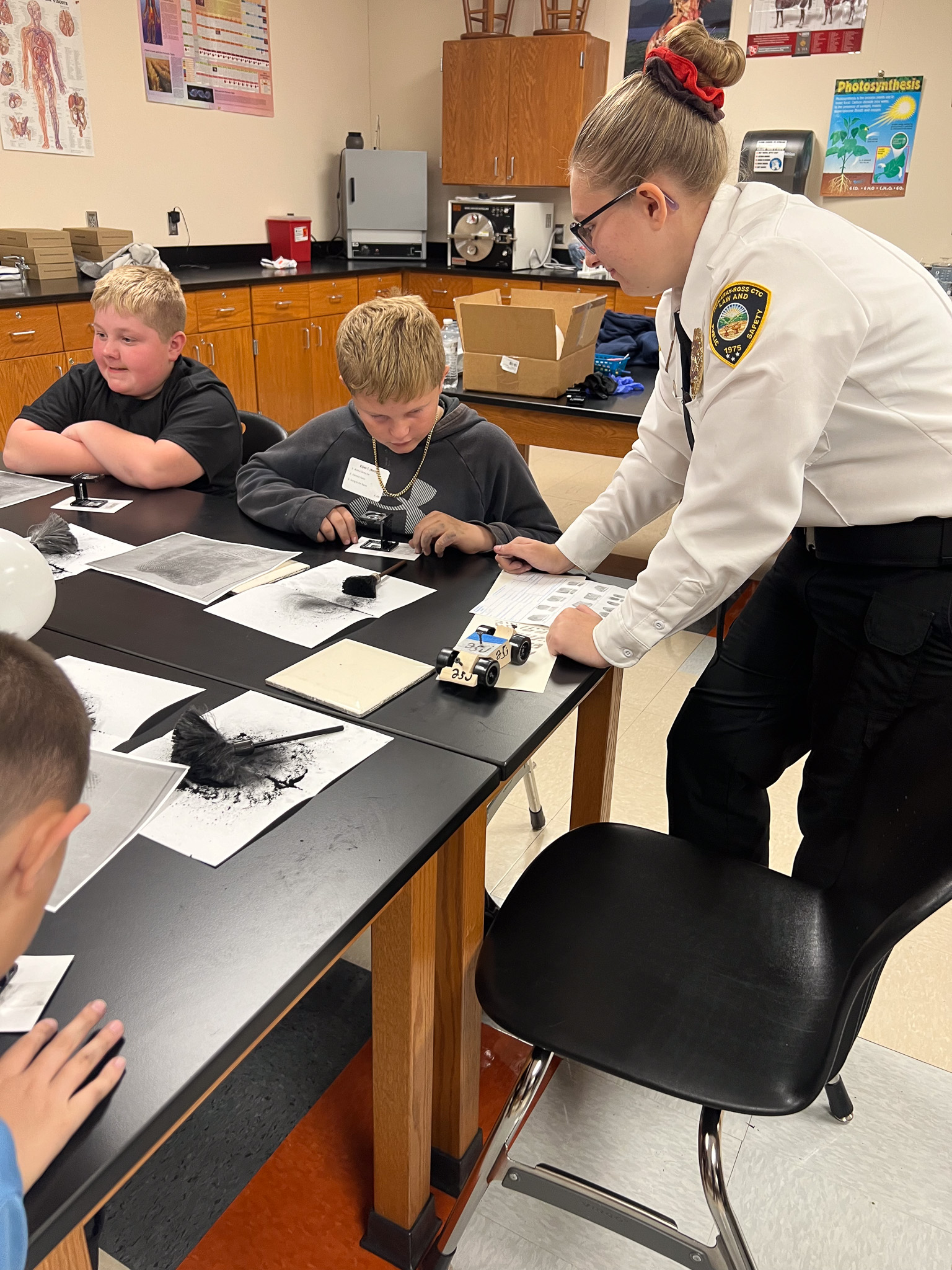A police officer assists a boy at a table in a classroom with a small car. Three other children look on.