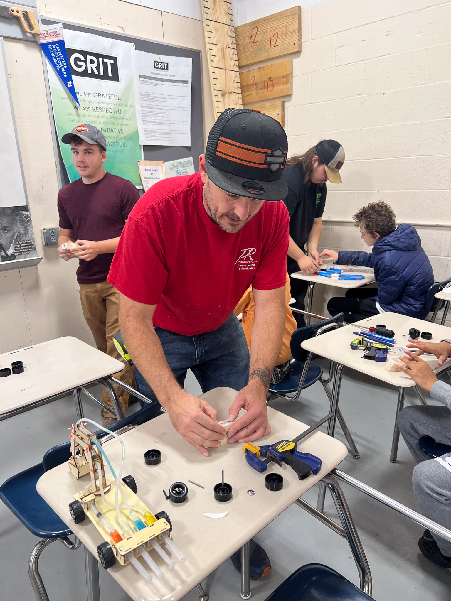 A group of people in a classroom, with one man in a red shirt assembling a toy car.