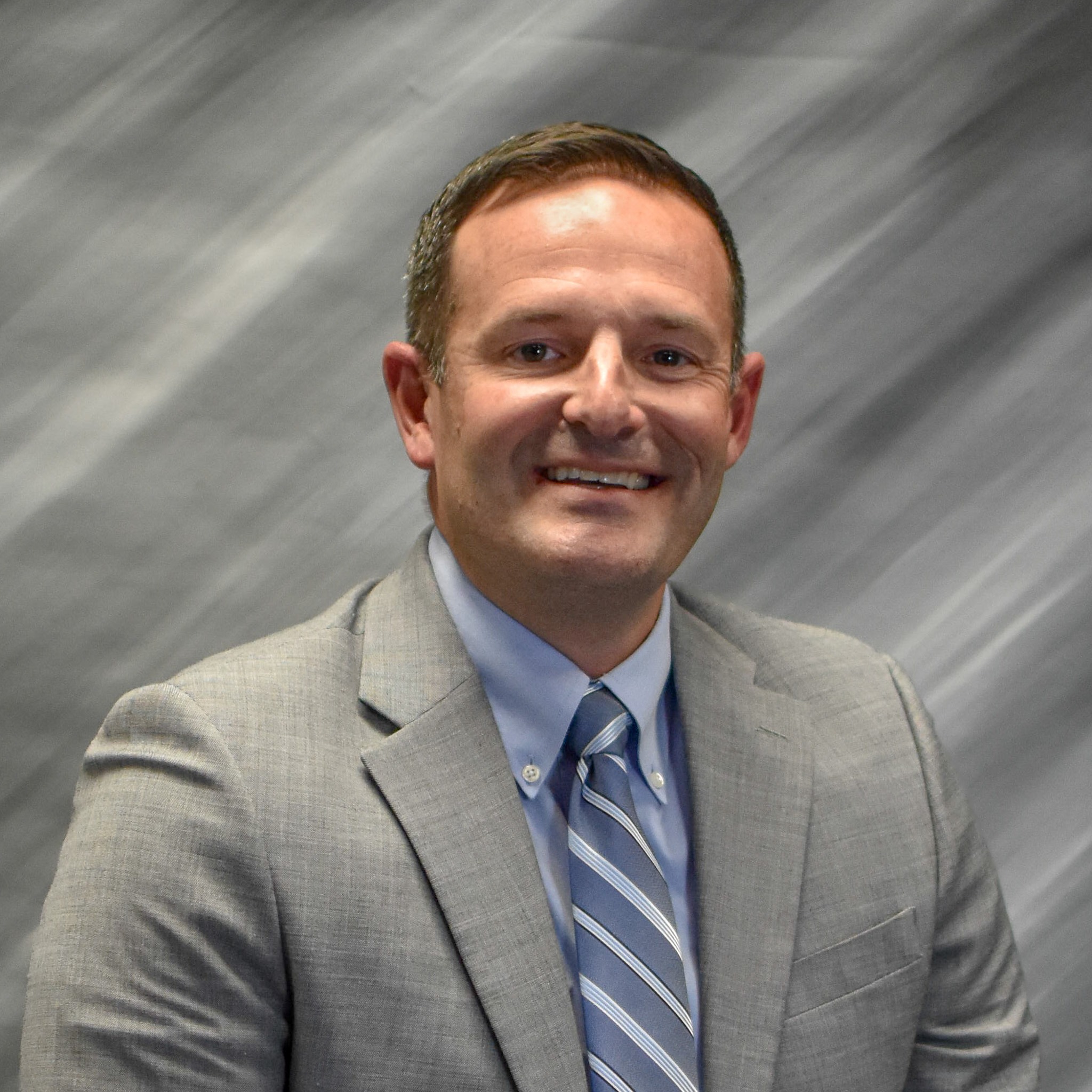 A man in a gray suit and blue tie stands in front of a gray gradient background.
