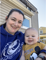 Amanda Pawlicki, a white woman with dark hair in a ponytail, is smiling into the camera with a jolly-looking baby and wearing a school hoodie