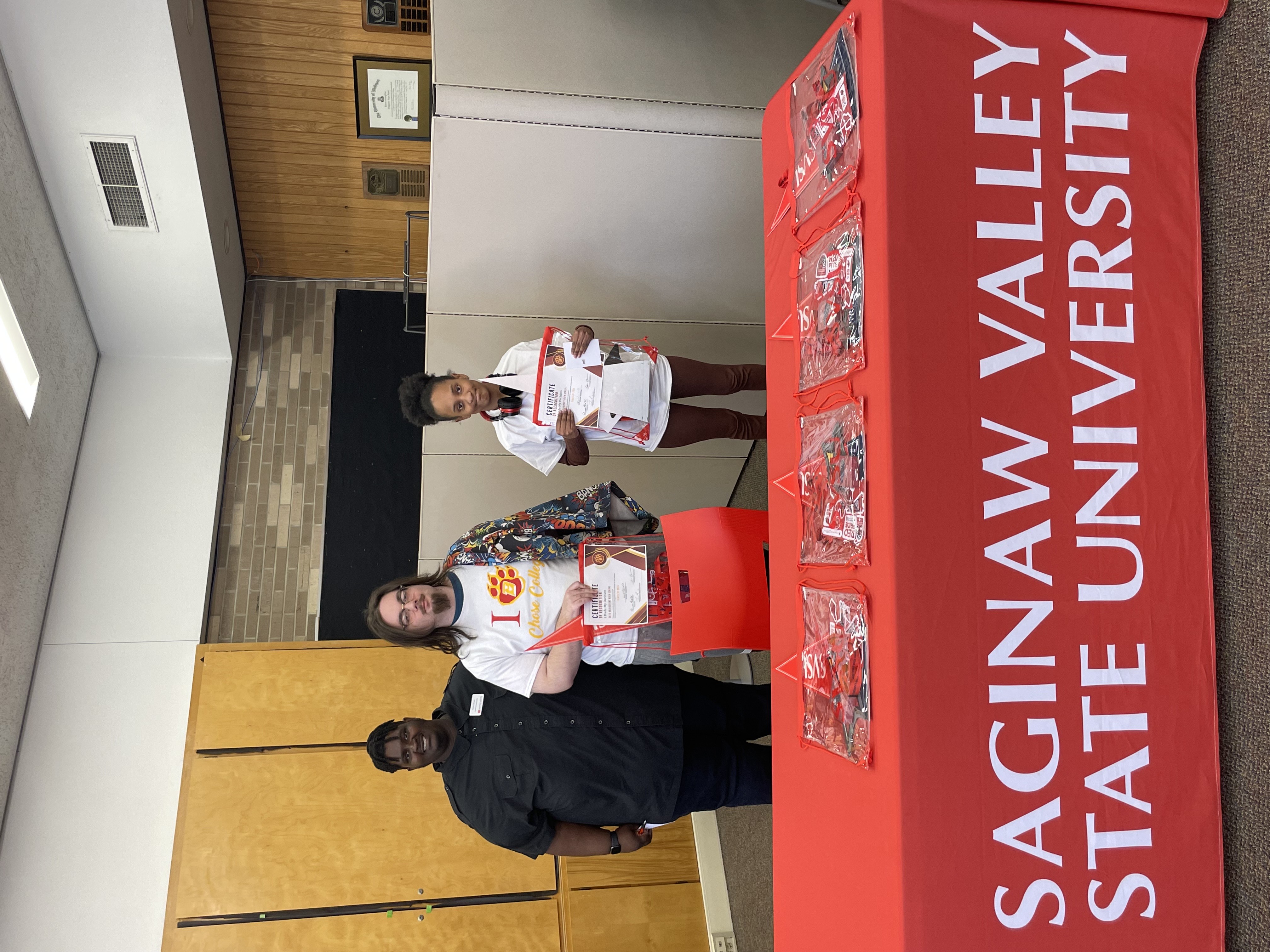 Three young people stand behind a table covered with a red tablecloth that reads "SAGINAW VALLEY STATE UNIVERSITY" in white letters. They are holding certificates and smiling. The person on the left has dark skin and dreadlocks and is wearing a black shirt. The person in the middle has light skin and long hair and is wearing a white t-shirt with a red "1" and a paw print design. The person on the right has medium skin and short curly hair and is wearing a white t-shirt and brown pants.