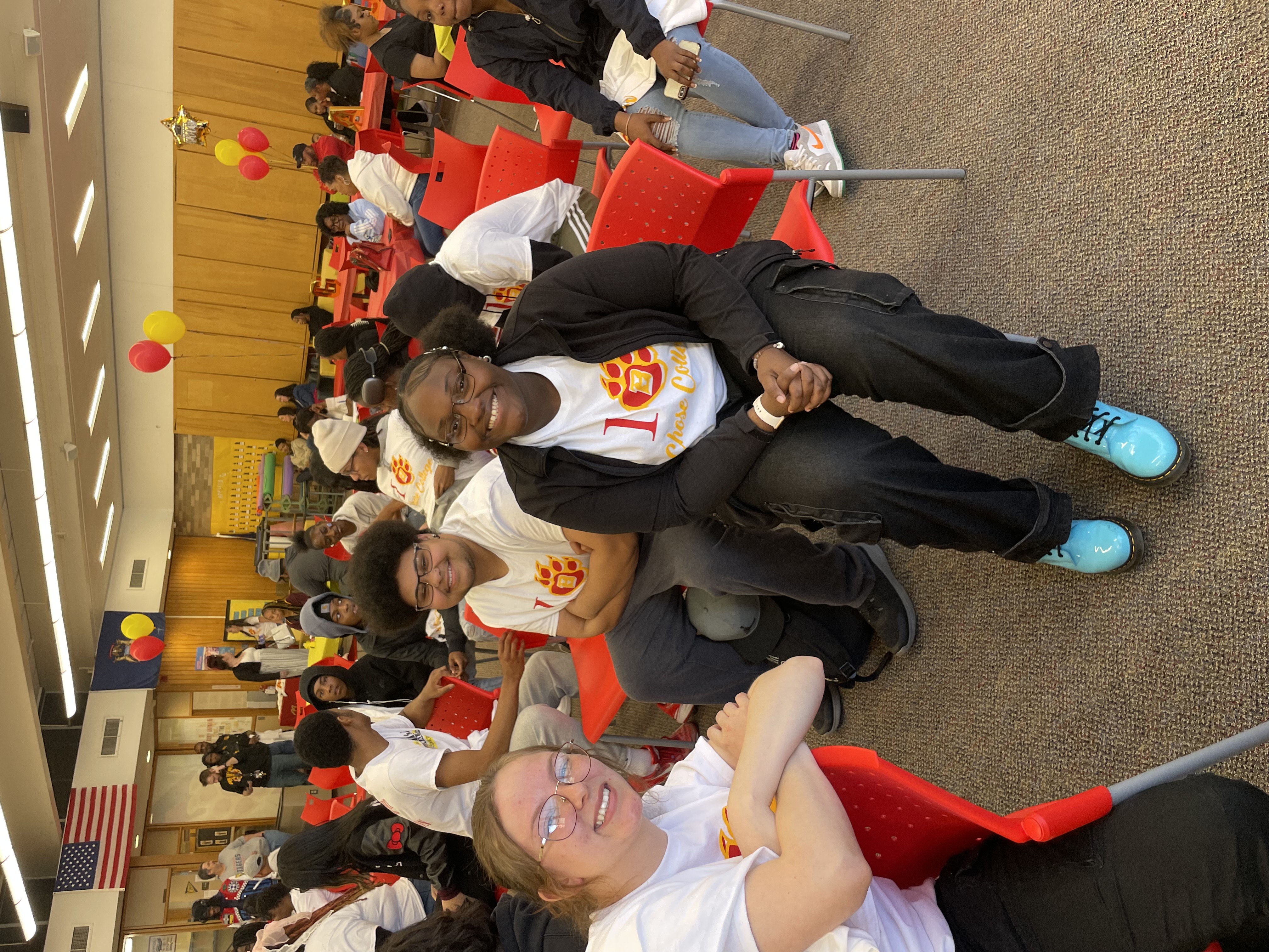A group of young people are seated in red chairs, some smiling at the camera. They are wearing white t-shirts with a red "1" and a paw print design. In the foreground, a young woman with glasses leans on the back of a chair, a young man with glasses sits with his arms crossed, and a young woman in a black jacket smiles directly at the camera. An American flag and another flag with yellow and blue are visible in the background, along with more red and yellow balloons.