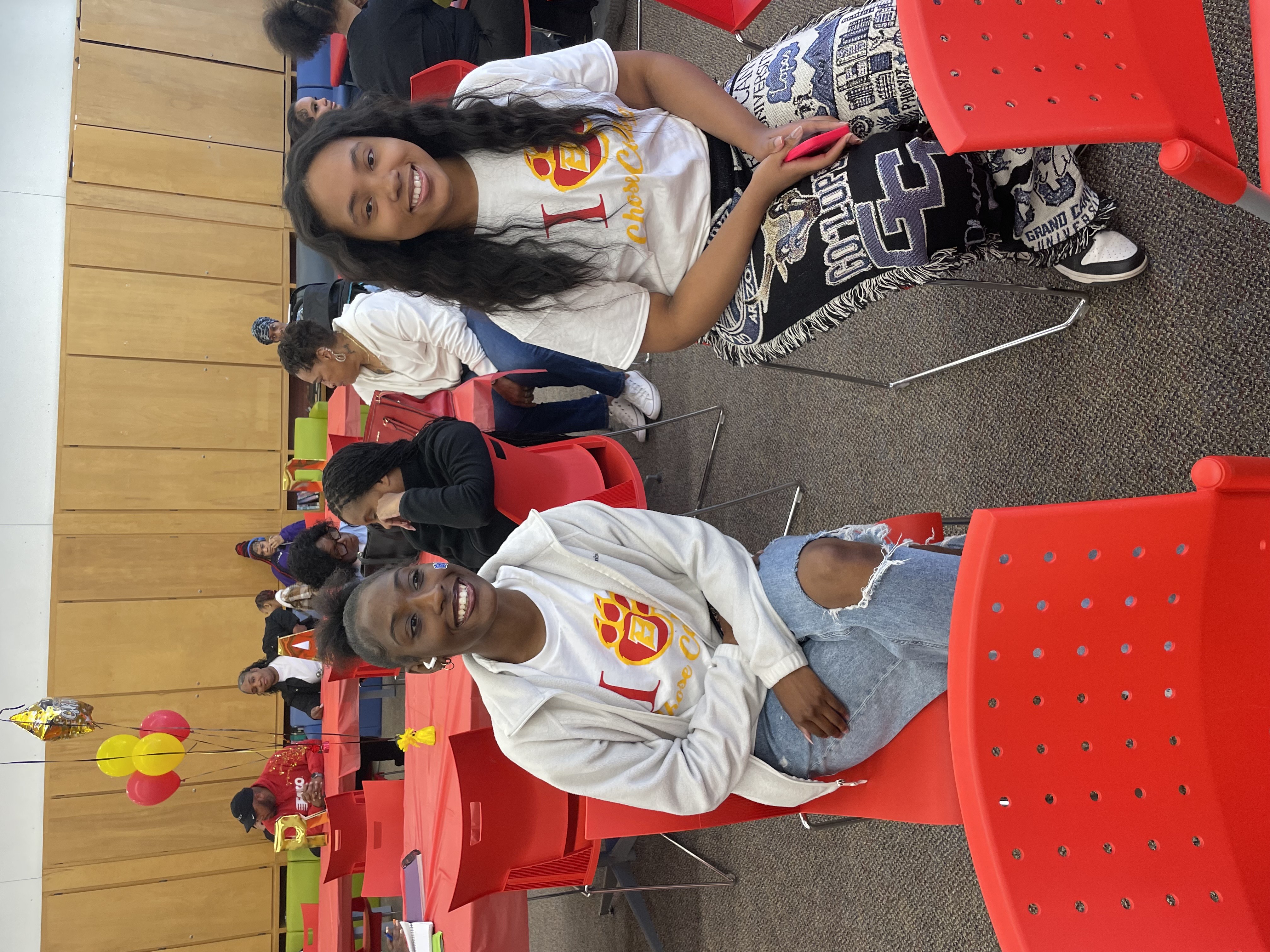 Two young women sit in red chairs, smiling at the camera. They are wearing matching white t-shirts with a red "1" and a paw print design. The woman on the left has her hair pulled back and is wearing a light-colored jacket and ripped jeans. The woman on the right has long, dark curly hair and is wearing patterned pants. Other people are seated at tables in the background.