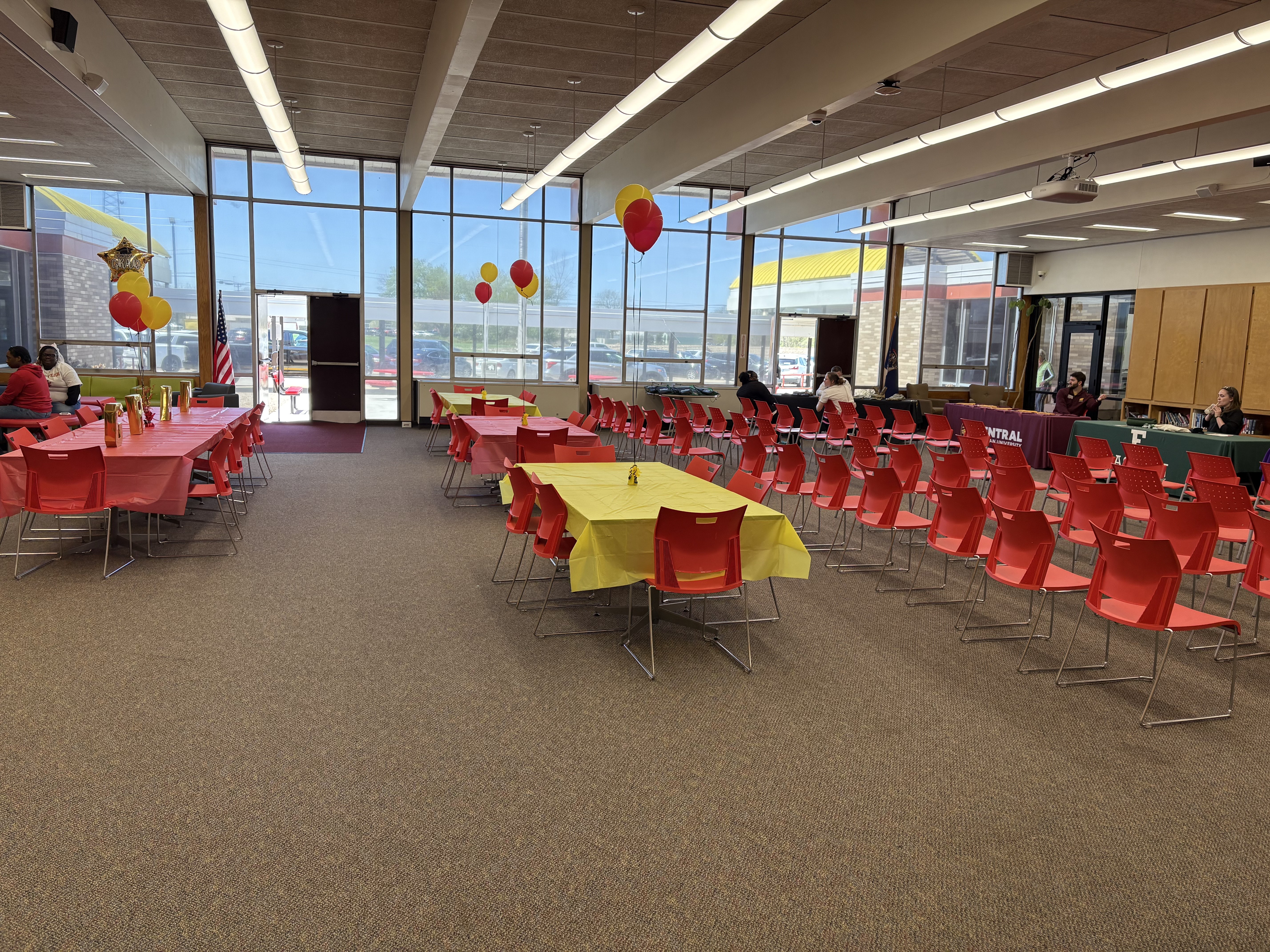 A brightly lit room, possibly a school cafeteria or large meeting space, is decorated with red and yellow balloons and streamers. Several long tables covered with red tablecloths and shorter tables with yellow tablecloths are arranged throughout the room, surrounded by red chairs. Some people are seated at the tables, and an American flag hangs in the background.