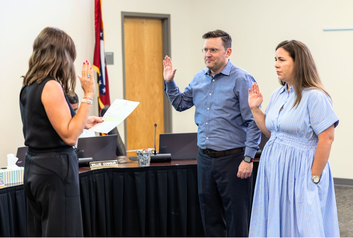 A man and woman raising their right hands looking at a woman holding up her right hand and reading a paper