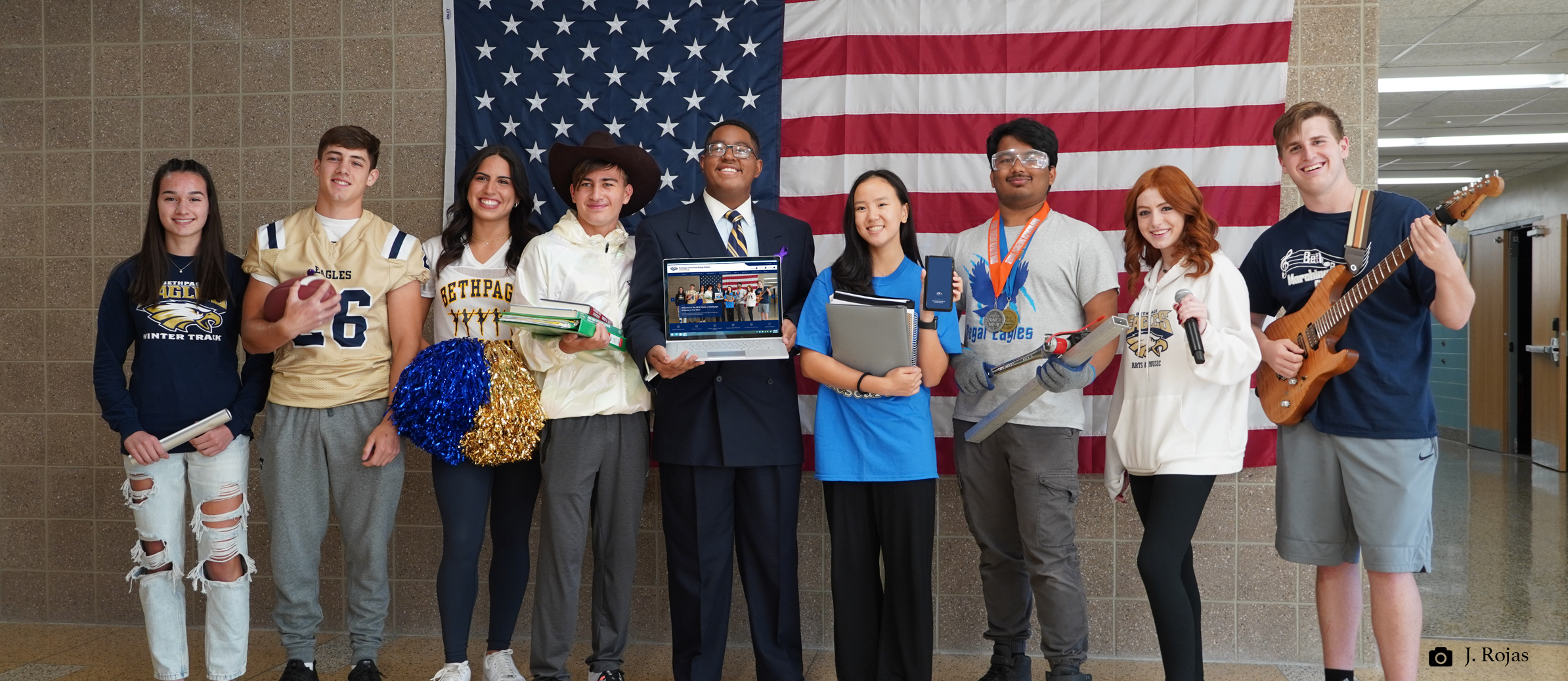 Students from various student activities standing in the school hallway in front of an american flag. Students are holding footballs, pom poms, laptops, phones, books, microphones, guitars