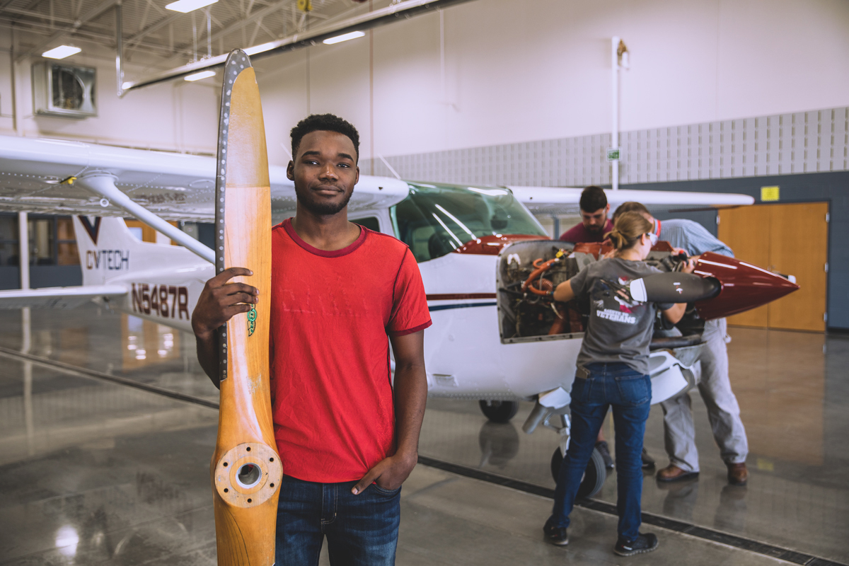 Young man holding airplane propeller while two students work on an airplane in the background.