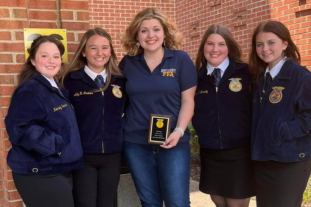 Four girls in FFA official dress pose for a photo in front of a brick building with FFA advisor Elizabeth Adams