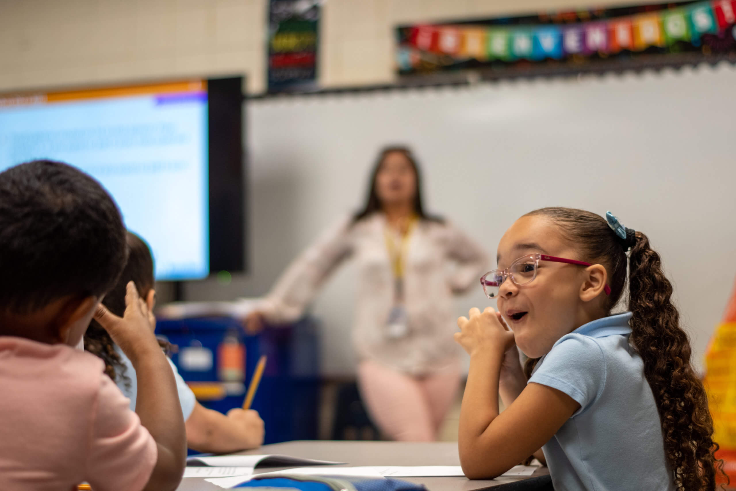 A young girl making an excited face towards something off screen with a teacher in the background