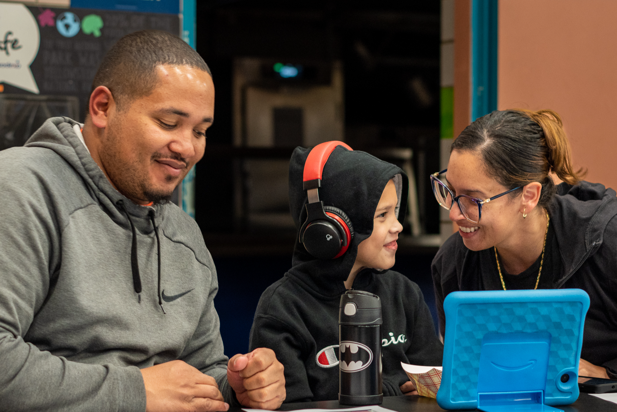 woman smiling at child wearing headphones in front of tablet while man looks on