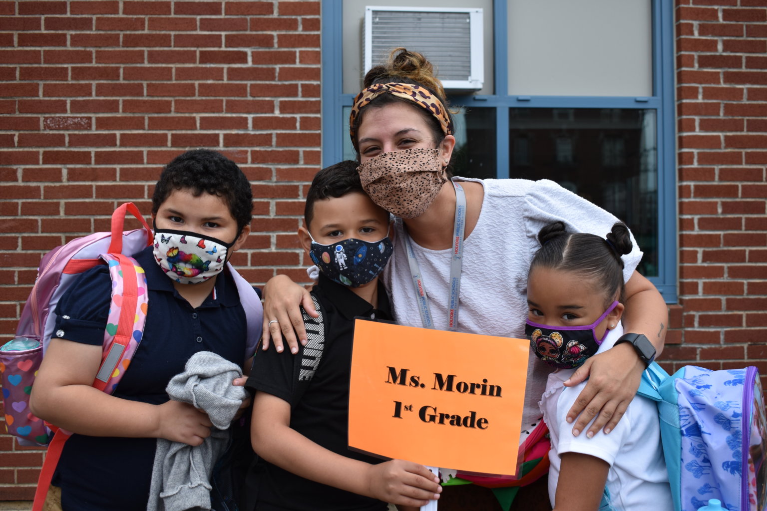 Teacher holding a sign that says Ms. Morin 1st Grade accompanied by three students
