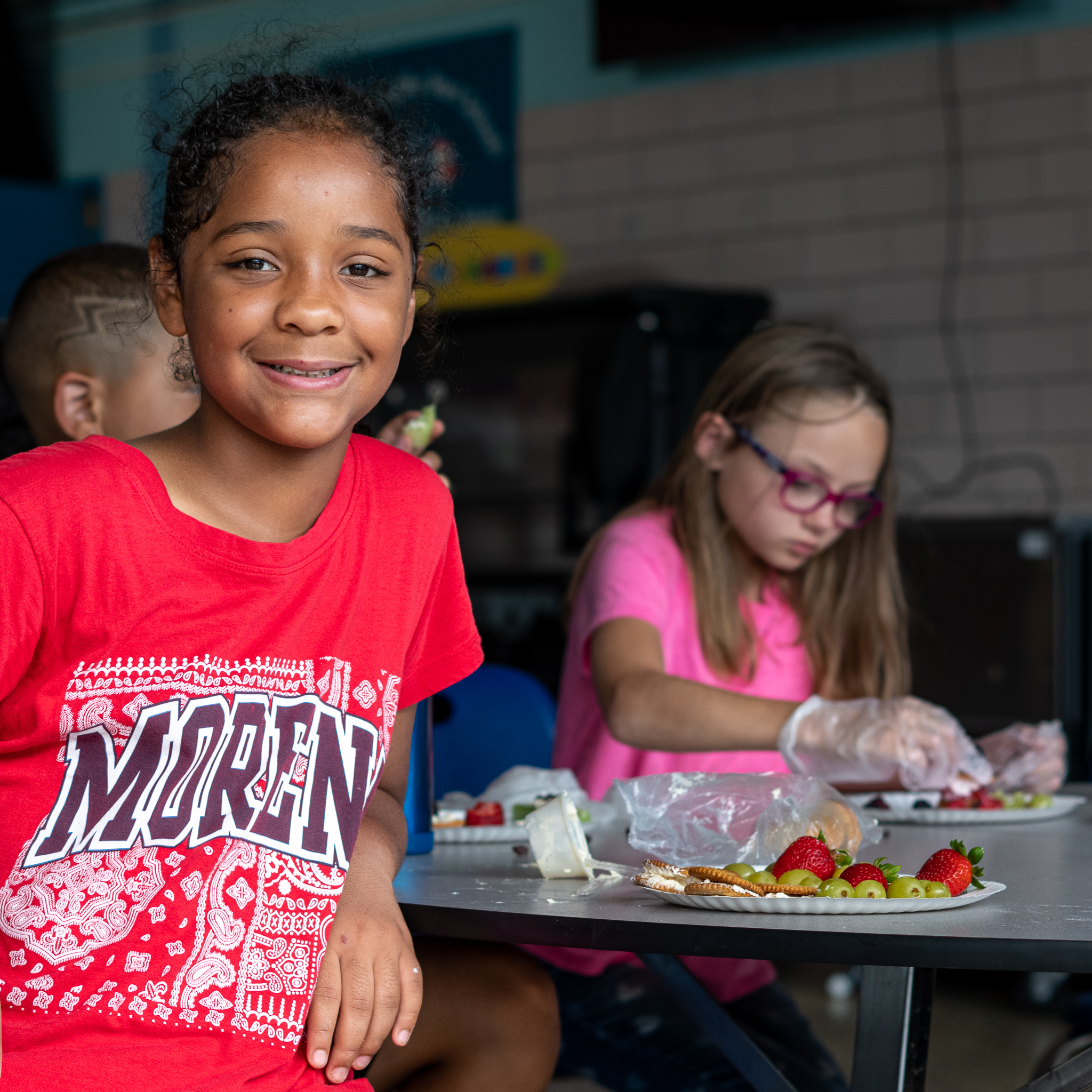 A smiling student next to a plate of fruit looking straight at the camera as other students behind her work on their plate.