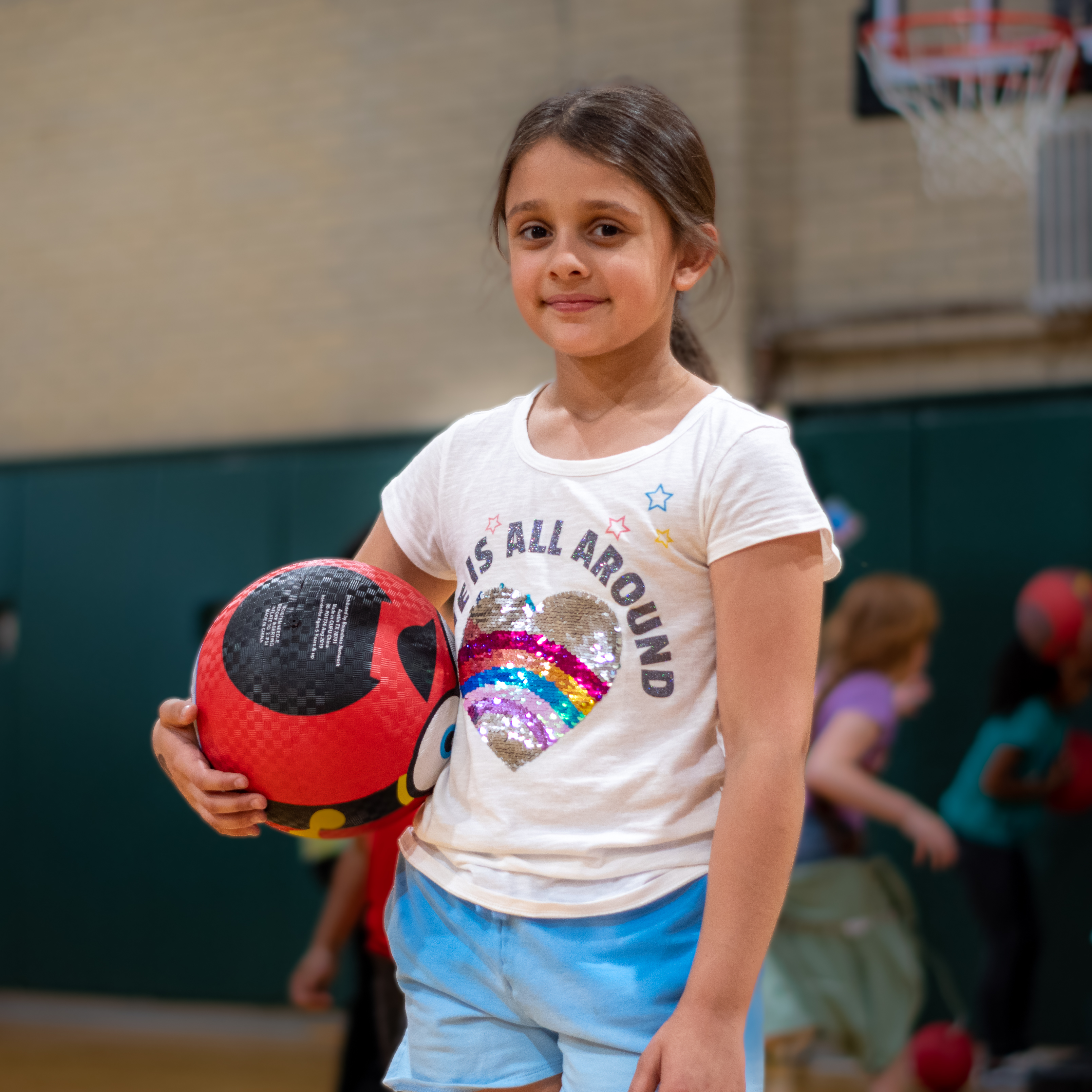 Student in gym class smiling as she holding a sports ball as other students in the background play. 
