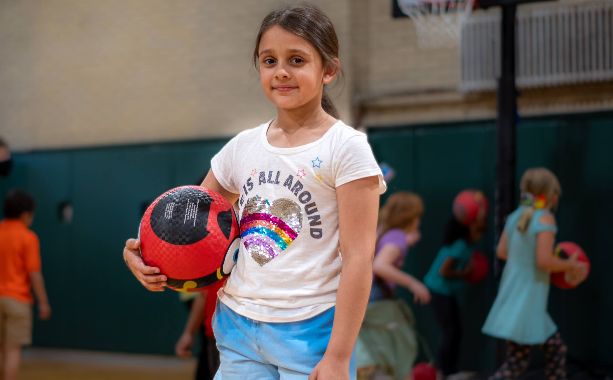 Student in gym class smiling as they hold a sports ball, as other students in the background play. 