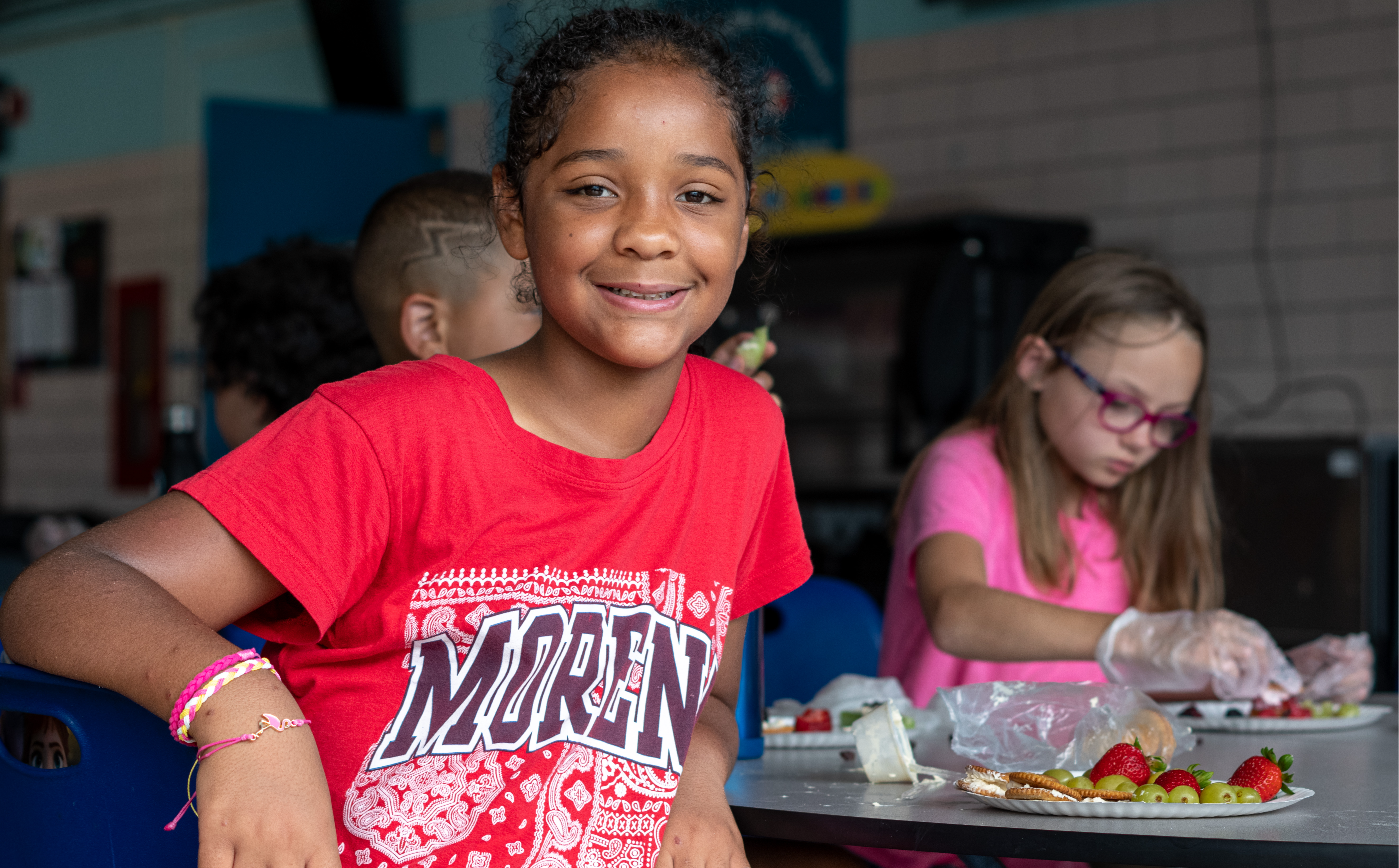 A smiling student next to a plate of fruit looking straight at the camera as other students behind them work on their own  plates.
