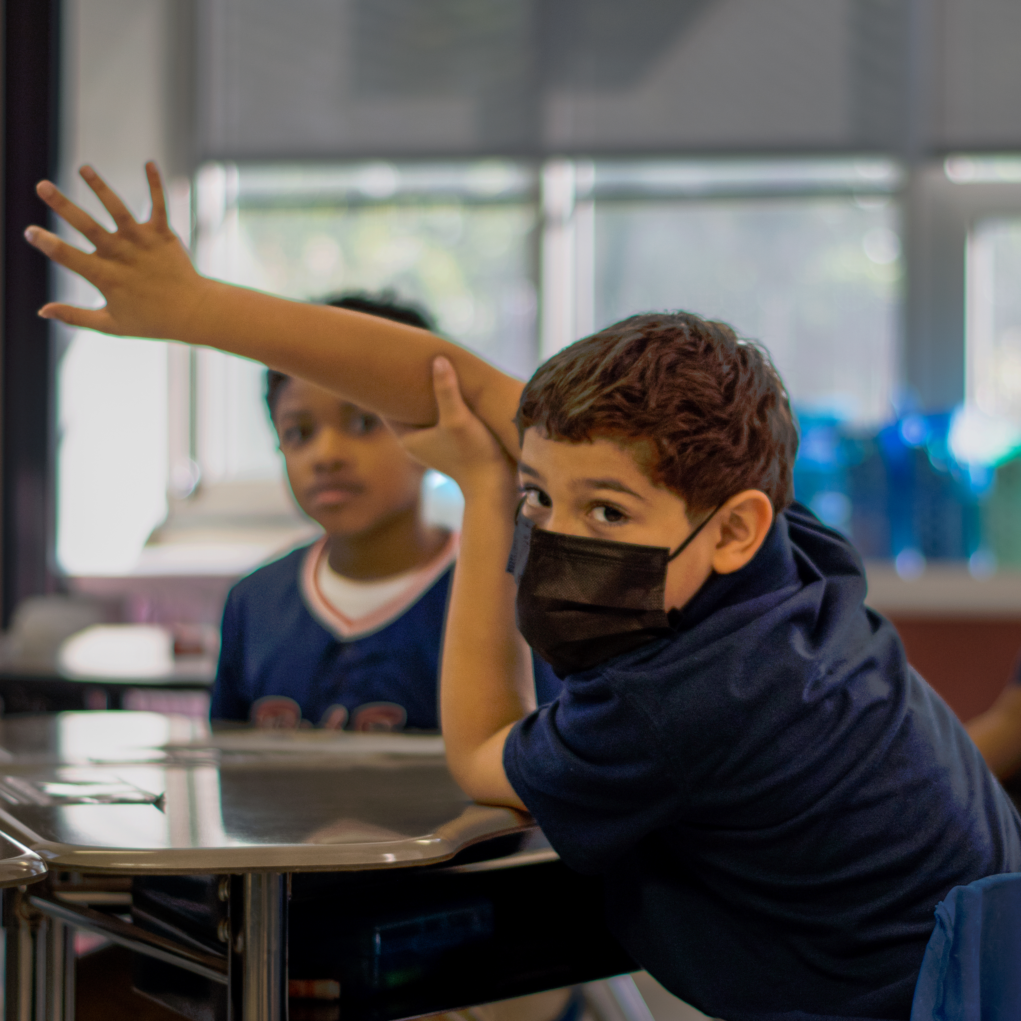 Two students  seated at desks, one raising their hand while looking into the camera as the other looks on.