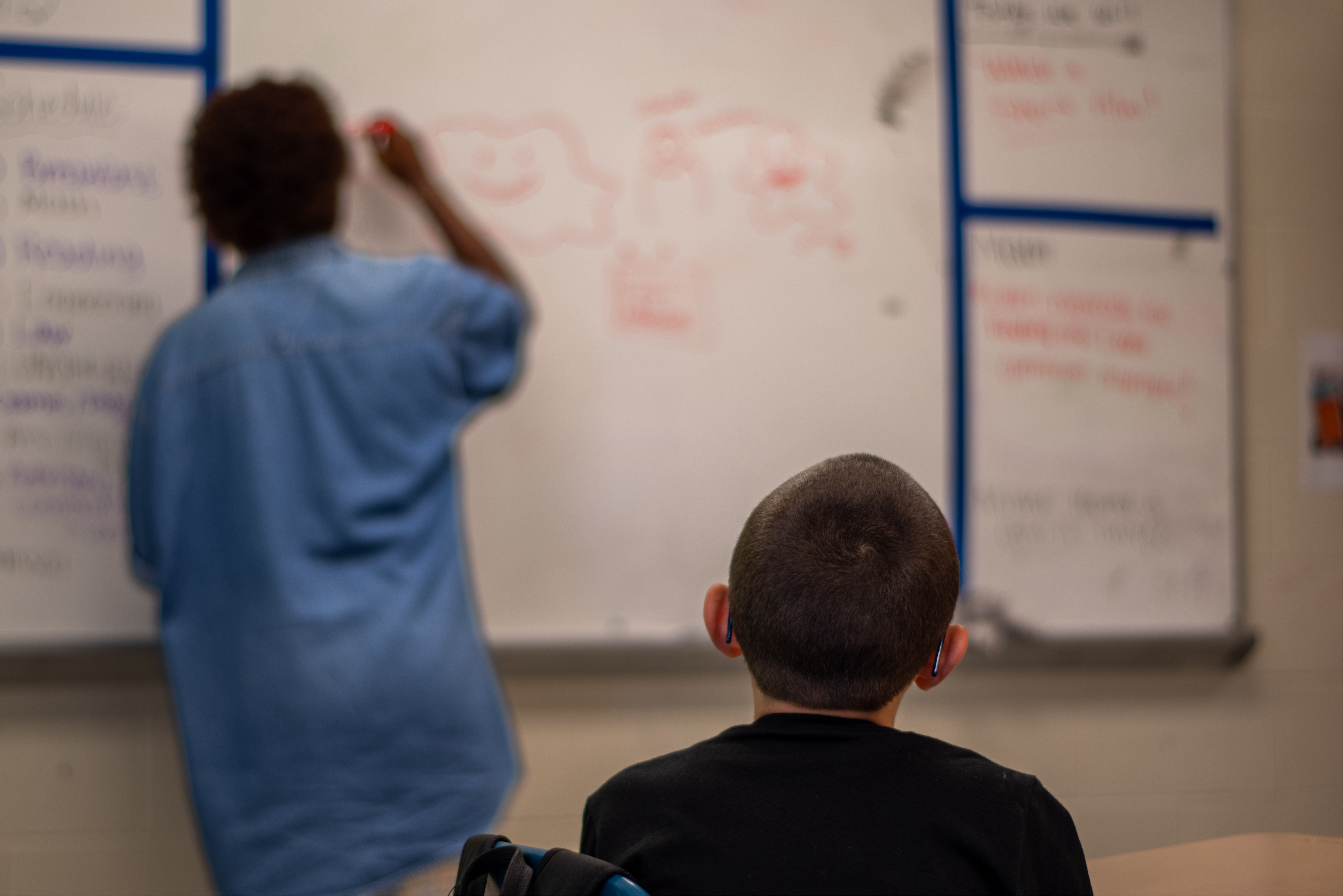 A student looking at the board as the teacher writes something in it