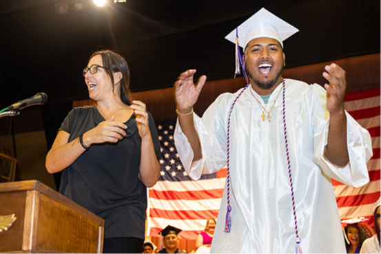Graduate wearing cap and gown smiling and clapping as teacher stands at podium