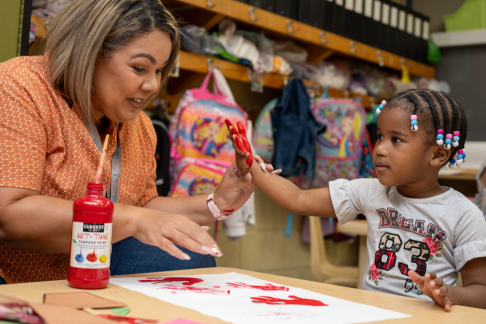 A teacher helping a student paint her hand for hand painting activity.