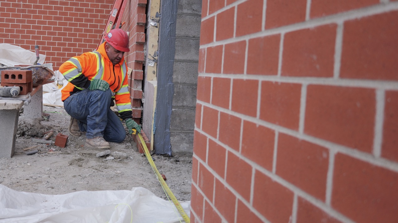 A worker measures close to the ground against an exterior wall