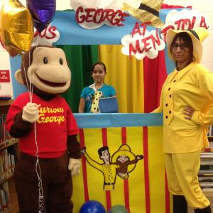 A  staff member dressed as the Man in the Yellow Hat posing with a student and a life size Curious George statue