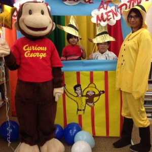 A  staff member dressed as the Man in the Yellow Hat posing with 2 students and a life size Curious George statue