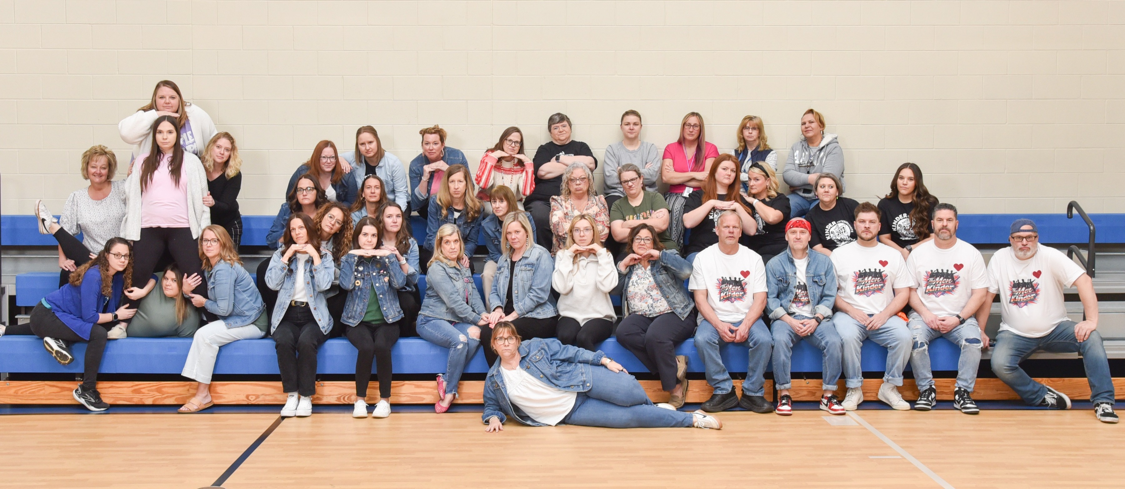 Group photo of people sitting and standing on bleachers in an indoor sports court, some wearing matching white shirts.