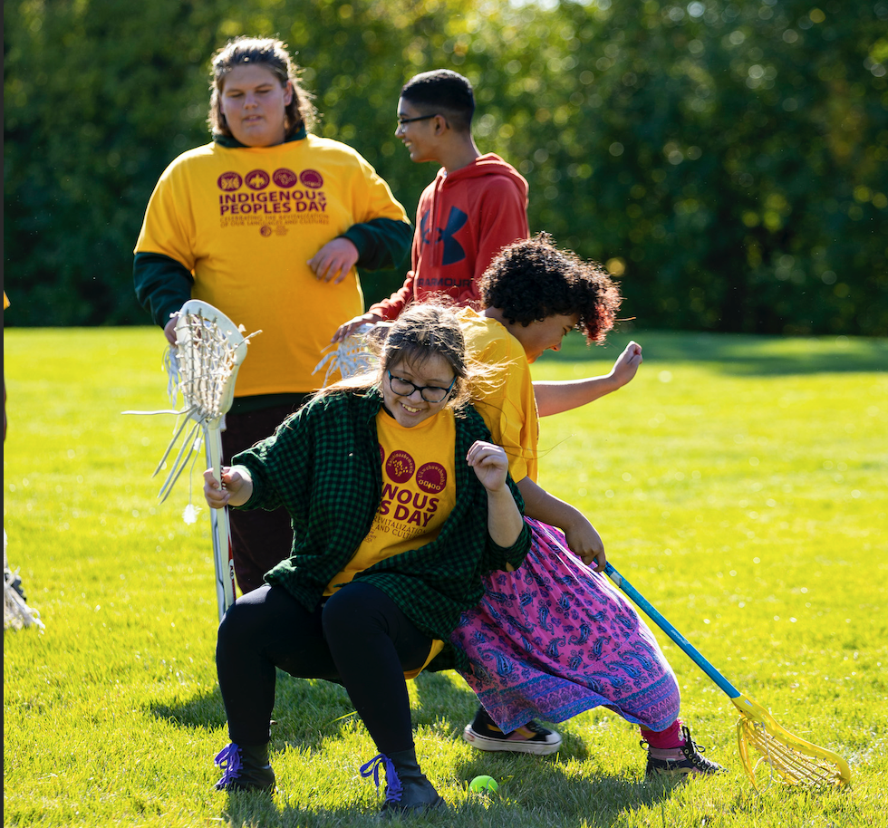 Four students wearing Indigenous Peoples Day t-shirts are playing lacrosse. Two girls in front are laughing.