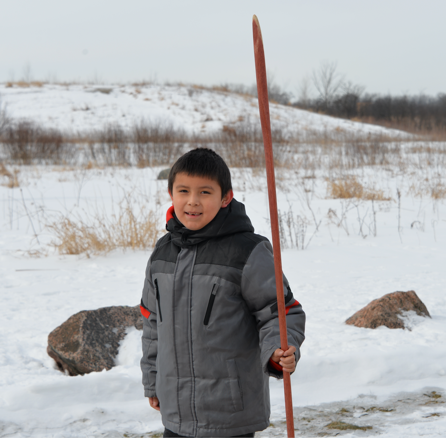 Boy wearing a gray coat is holding a snow snake wooden pole outside. There is a snow hill,  tall grasses and large rocks in the background.