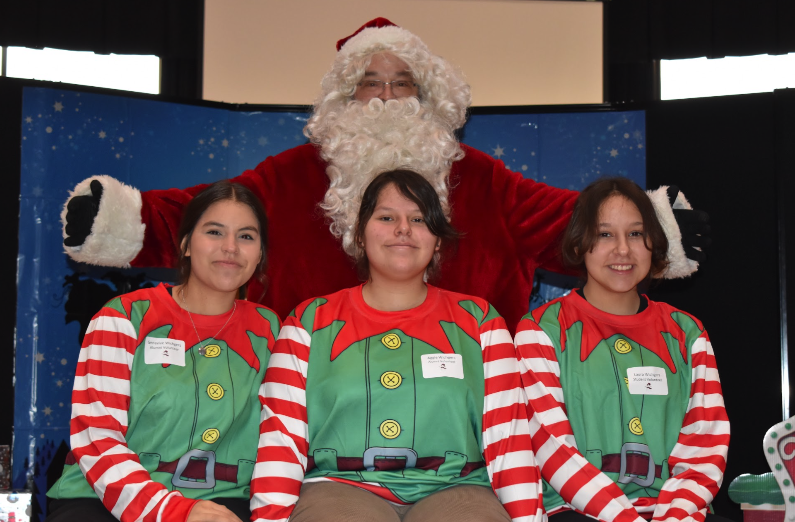 Three female students wearing green and red striped elf outfits with Santa behind them.
