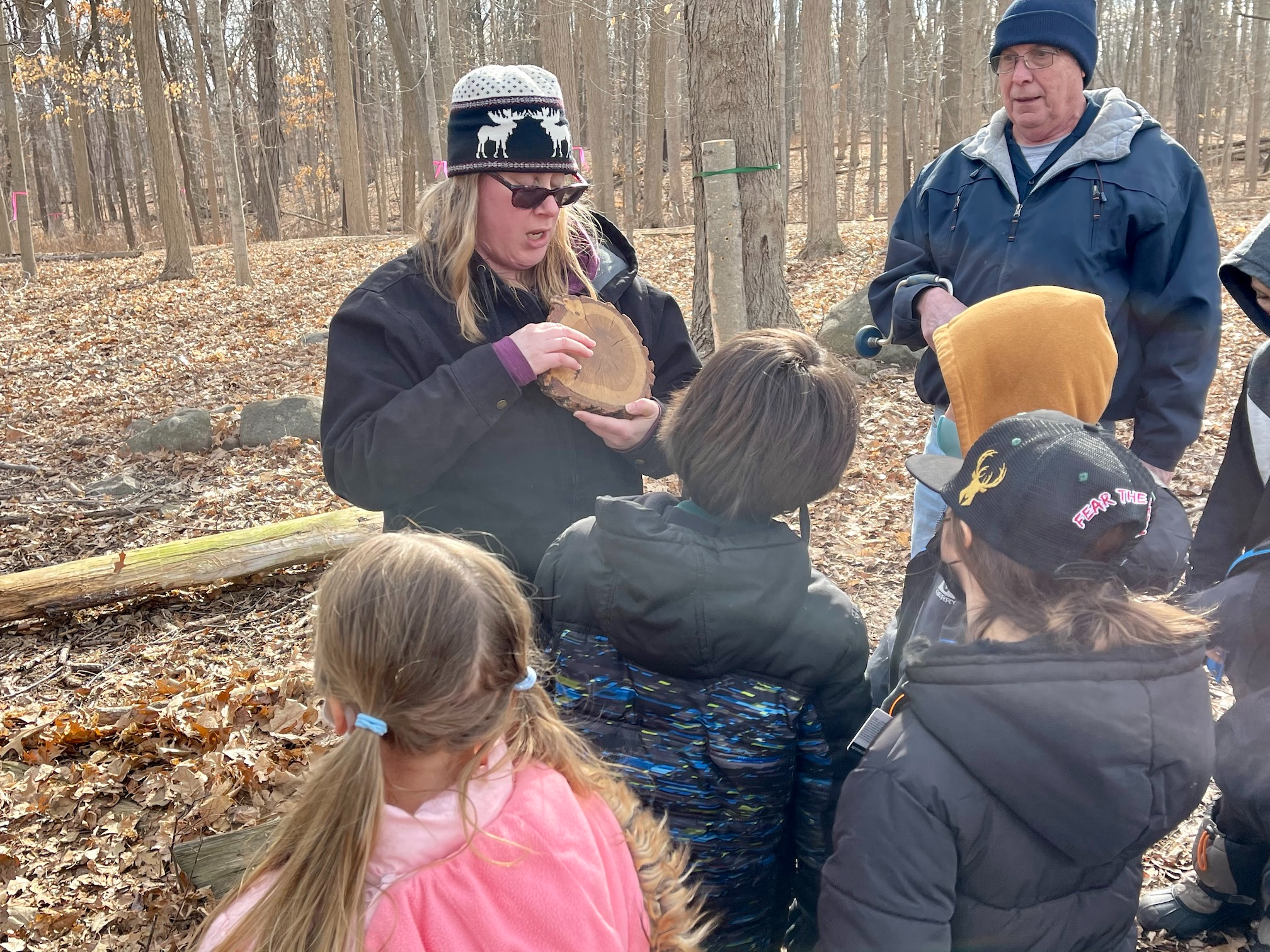Two adults outside in the woods with a group of students. They are showing them rings in a tree trunk to teach them about how to tell the age of a maple tree.