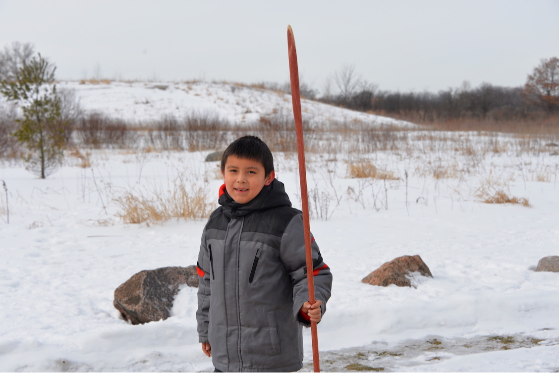 Little boy outside wearing his winter coat, holding a snow snake pole with snow all around him.