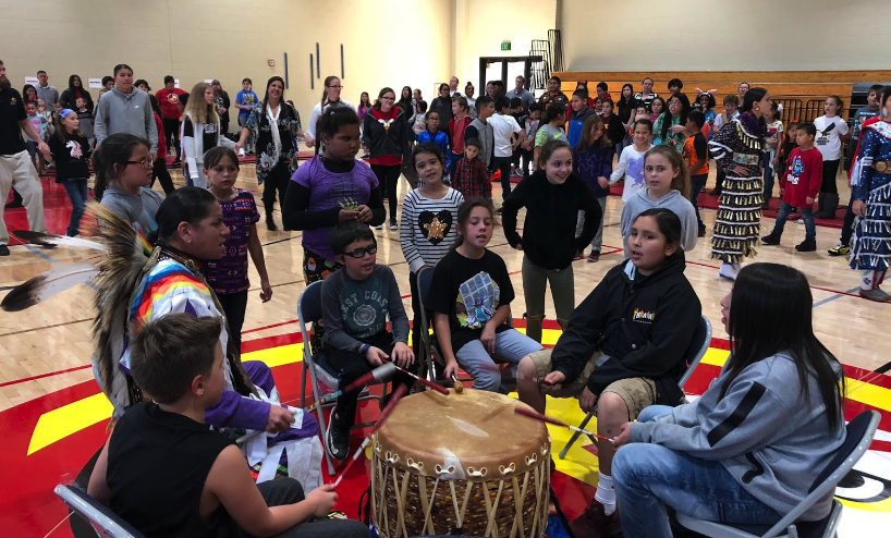 Students inside the gym around the pow wow drum with many students in the background.