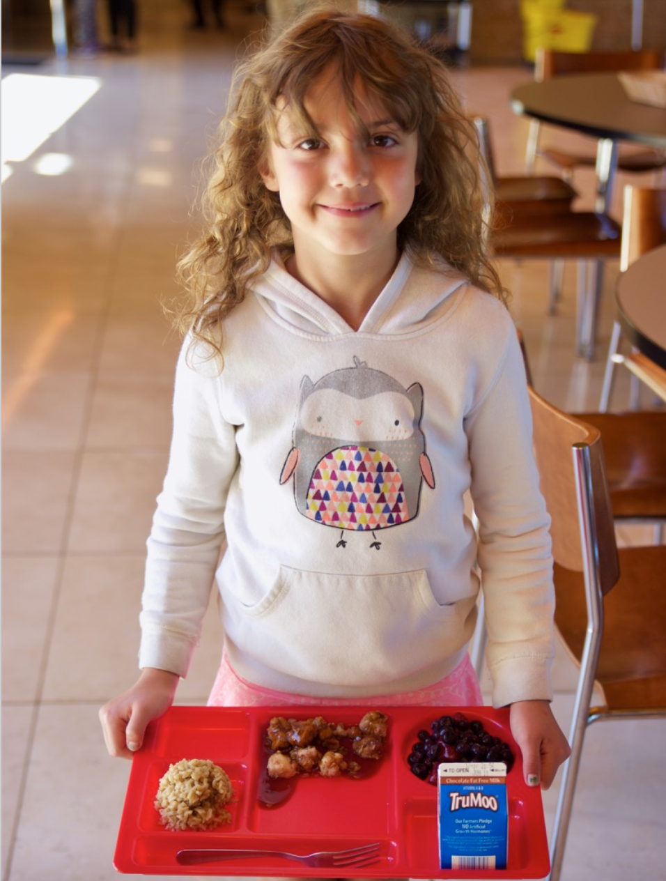 A young girl wearing a white sweatshirt with an owl on it, is smiling at the camera as she holds her red lunch tray with rice, meat, fruit and a carton of milk.  