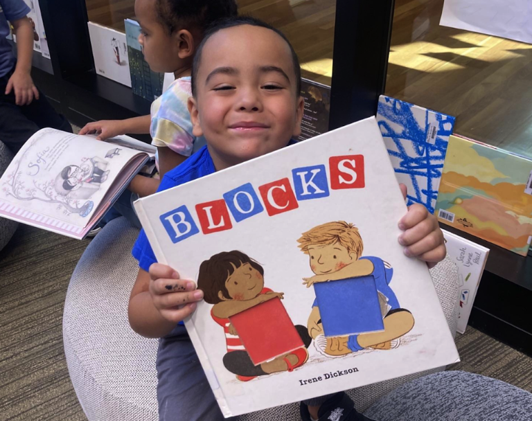 A young boy holding up his book titled, "Blocks". He is smiling and there is another child in the background.