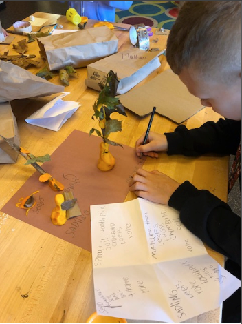 A boy student is working on his PBL project that has leaves. He is writing something on his project; his handwritten notes are also in the photo.