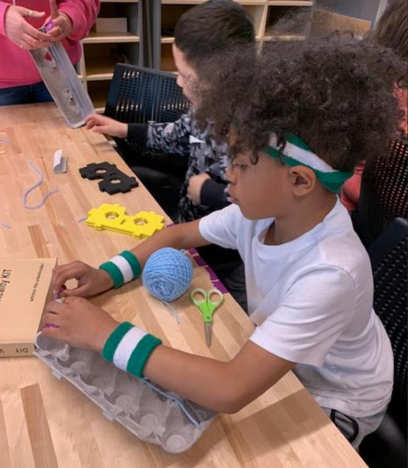 Young student wearing a white t-shirt, and matching green and white sweatbands on his head and writsts, is making something with a cardboard egg carton, blue yarn, and scissors. A second student is in the background working with black and yellow sytrofoam pices. A teacher's hands and pink top is also in the background.