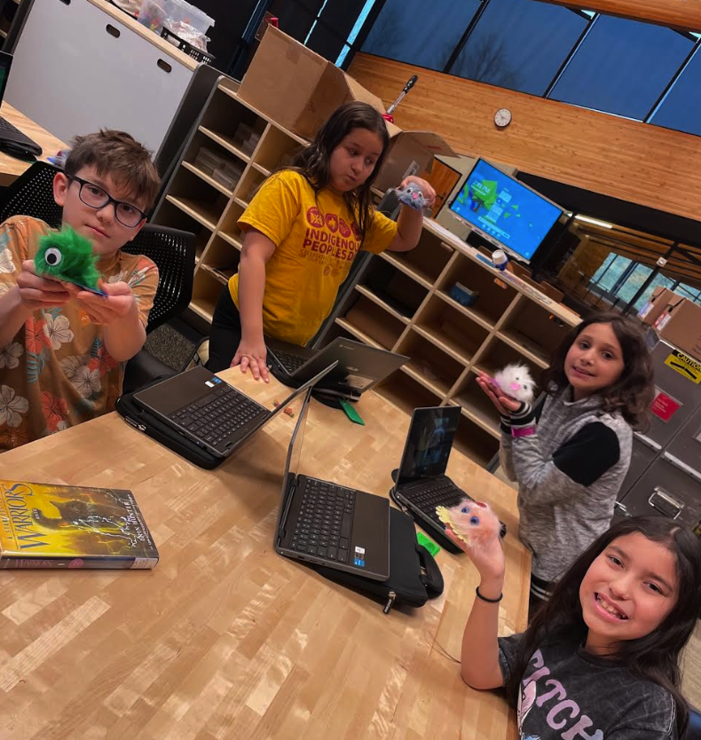 Four students are holding up their fuzzy monster creatures they made in Makerspace. They have their laptops opened up in front of them; they are seated at a wooden table.