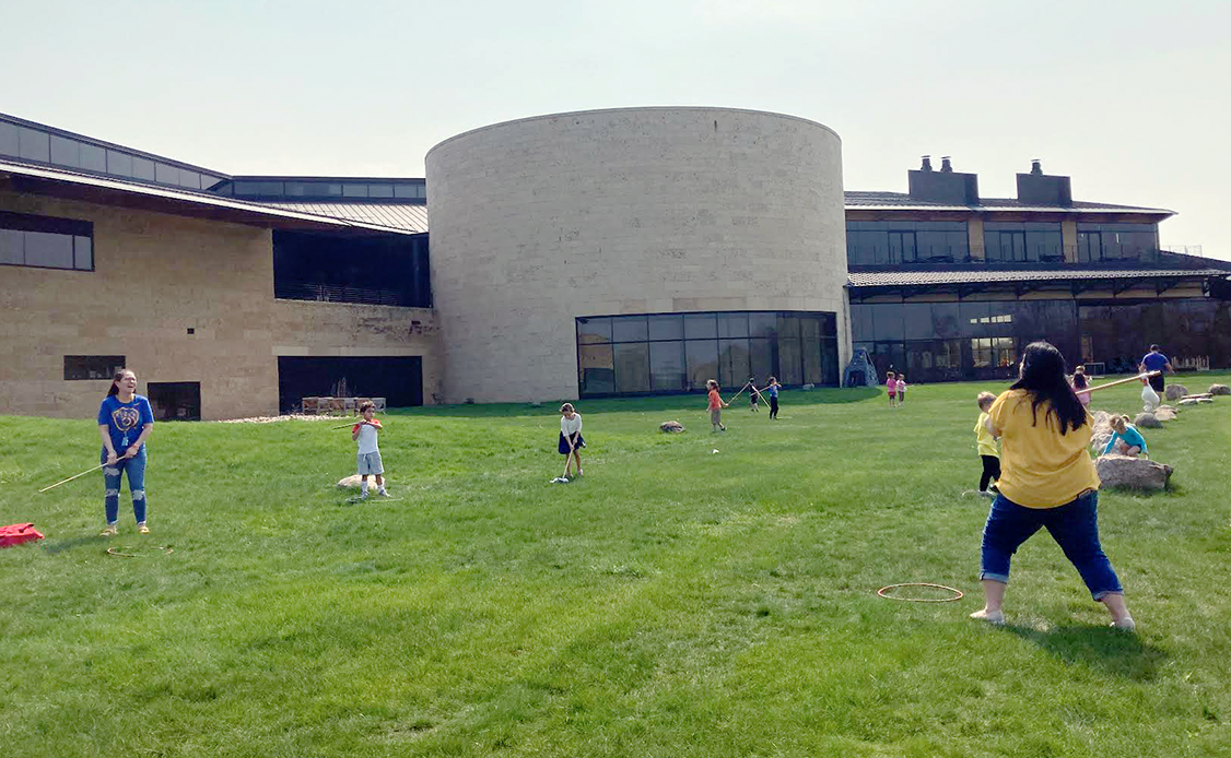 Twelve students are outside on the grass; the school building is behind them. They are learning how to play double ball.