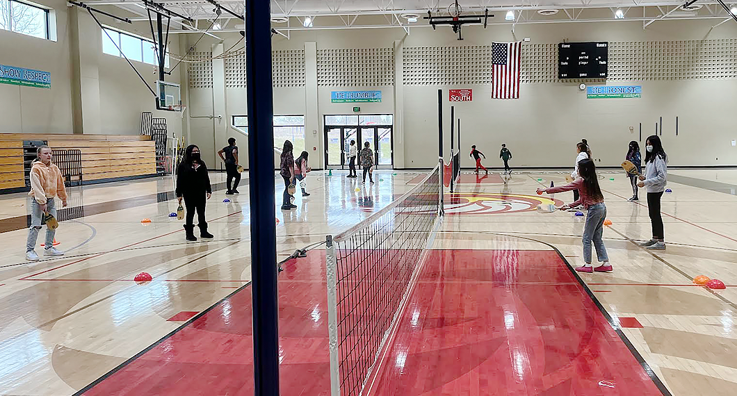 14 students are inside the gym playing paddle ball. There is a low net dividing the gym floor and some of the students are wearing face masks.