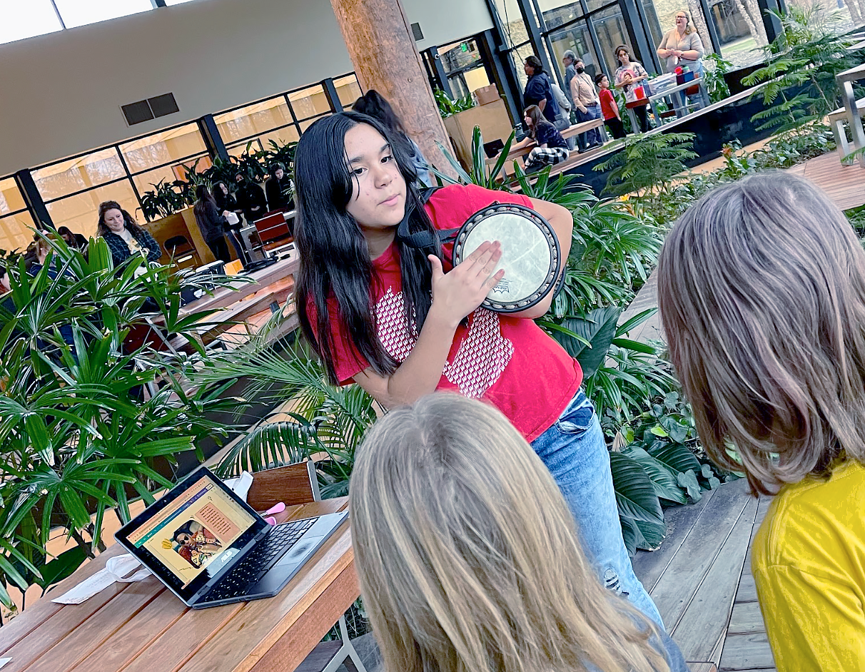 Female student wearing a red t-shirt, is holding and playing a bongo drum for two blond students in the foreground. A laptop computer is open. They are inside with plants all around them and teachers and students are far away in the background.