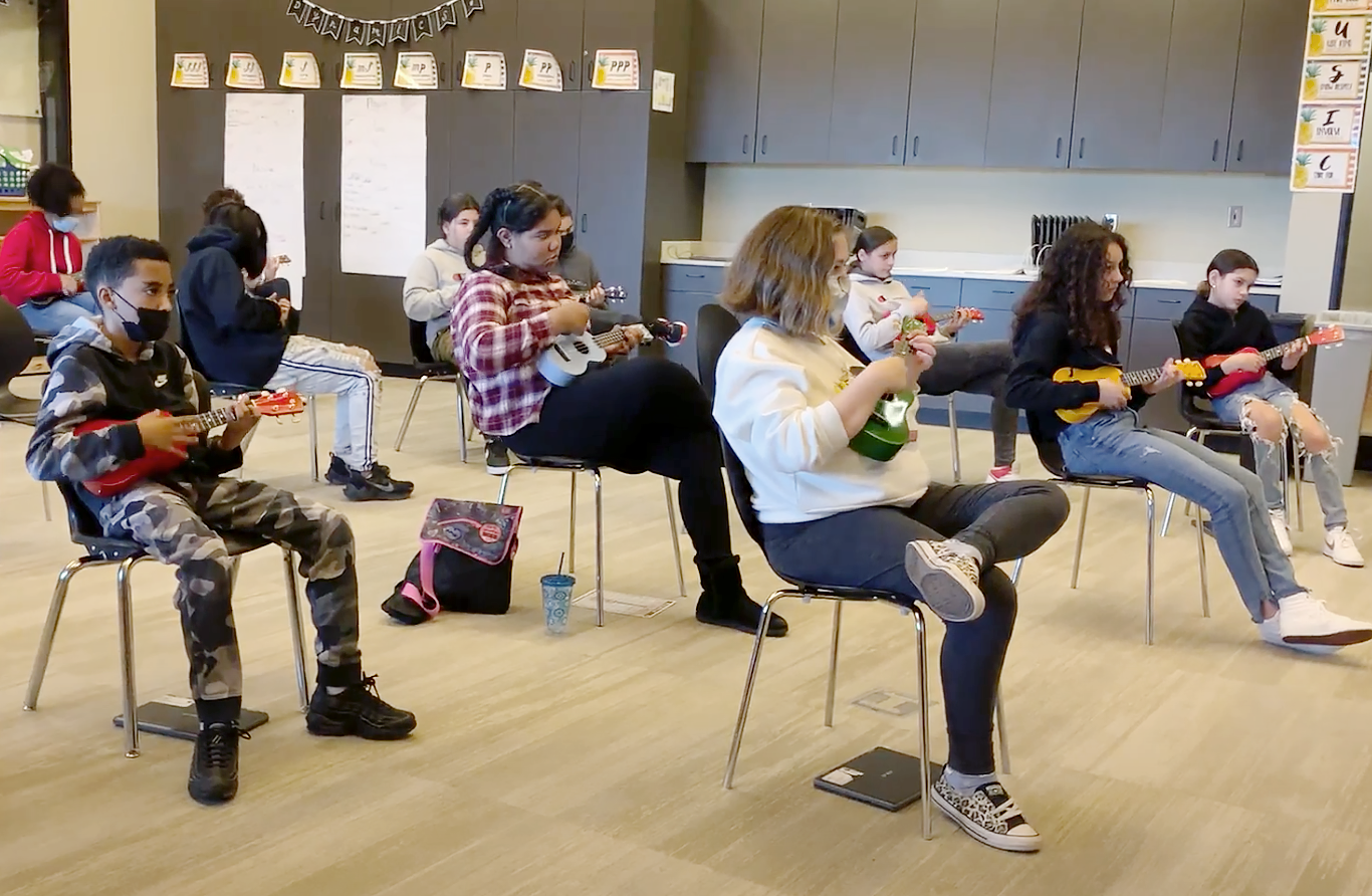 Classroom of students seated on chairs, playing ukuleles. Two are wearing facemasks.