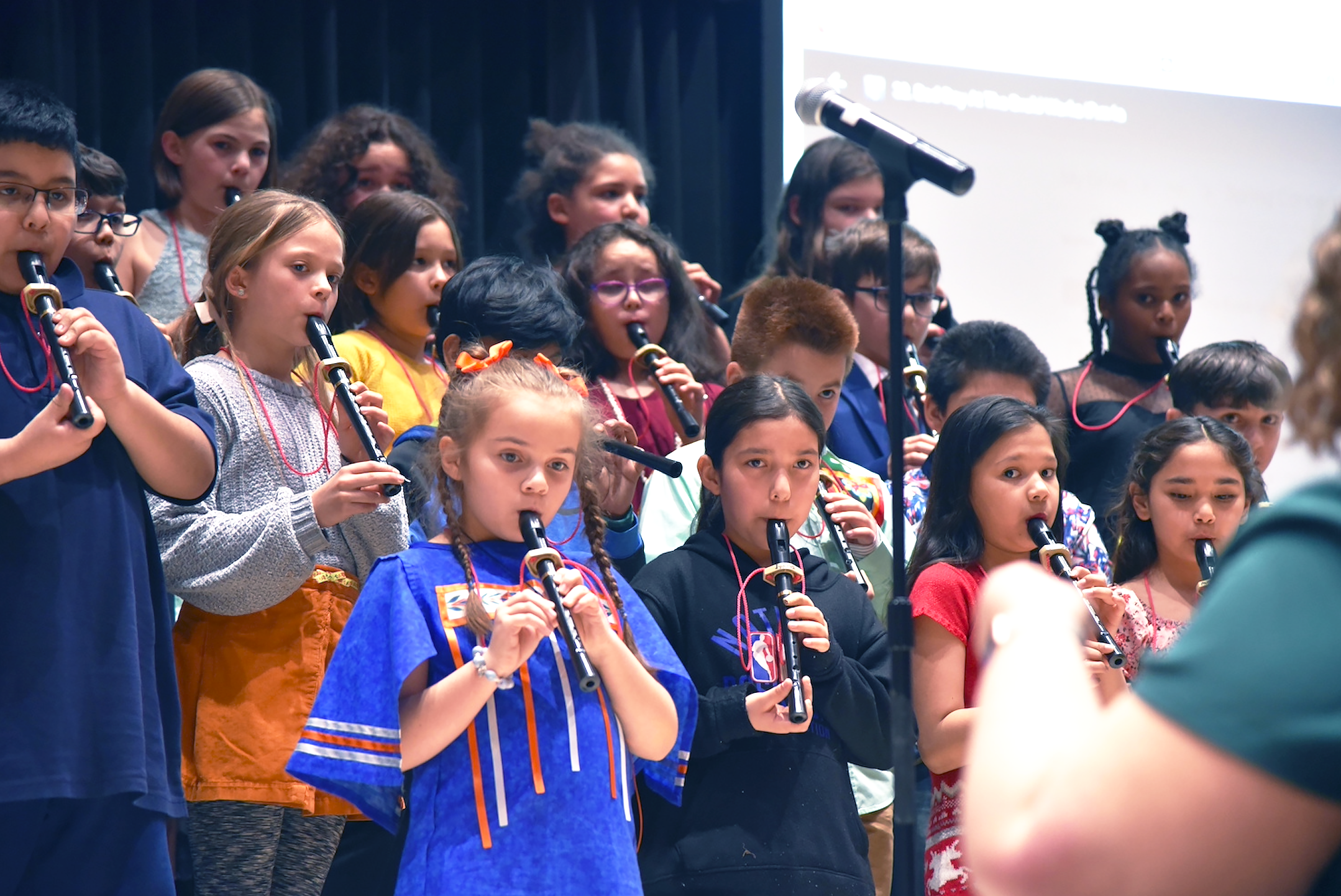 Group of students playing recorders at a concert; teacher is on the right, conducting them.