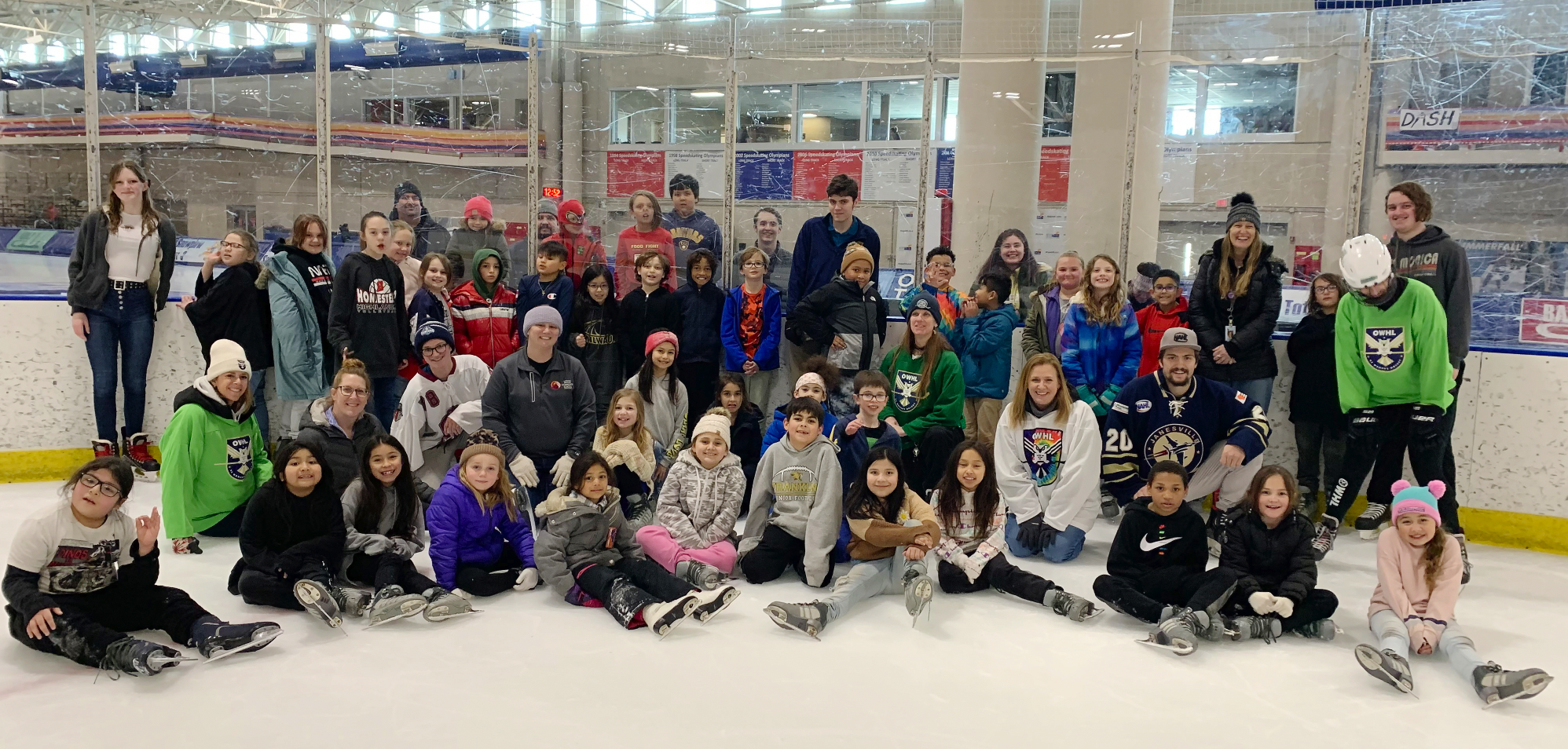 Large group of ICS students wearing hats, skates and smiles, as they sit on the ice at the Pettit Center with teachers and chaperones.