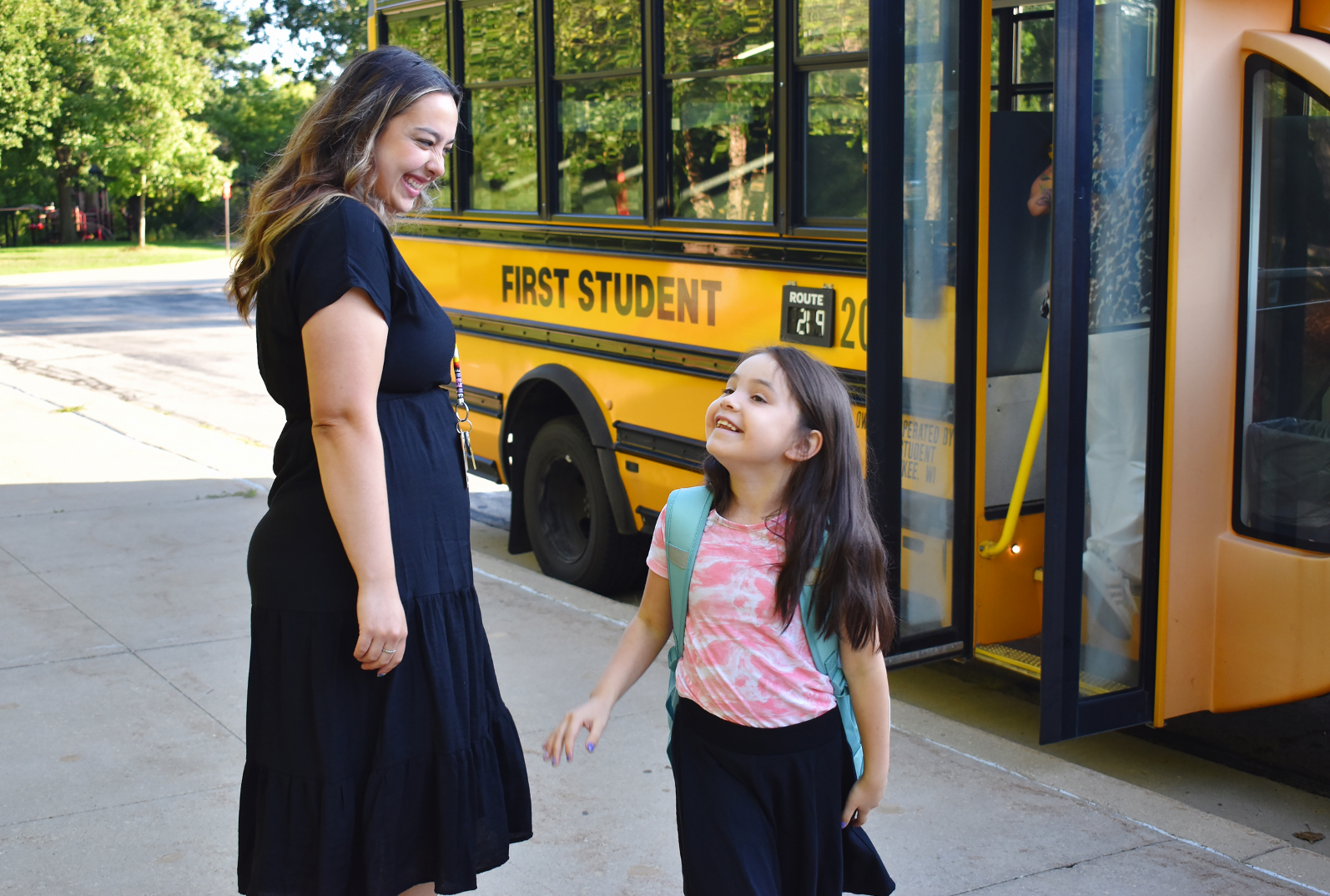 Female teacher wearing a black dress is smiling at a young girl student, who acknowledges her as she walks by. The school bus is in the background.