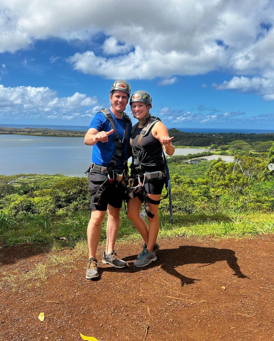 Husband and wife on hillside overlooking blue water bay; they are wearing gray helmets, safety harnesses, shorts, tennis shoes, and giving the "hang ten" sign with their hands.