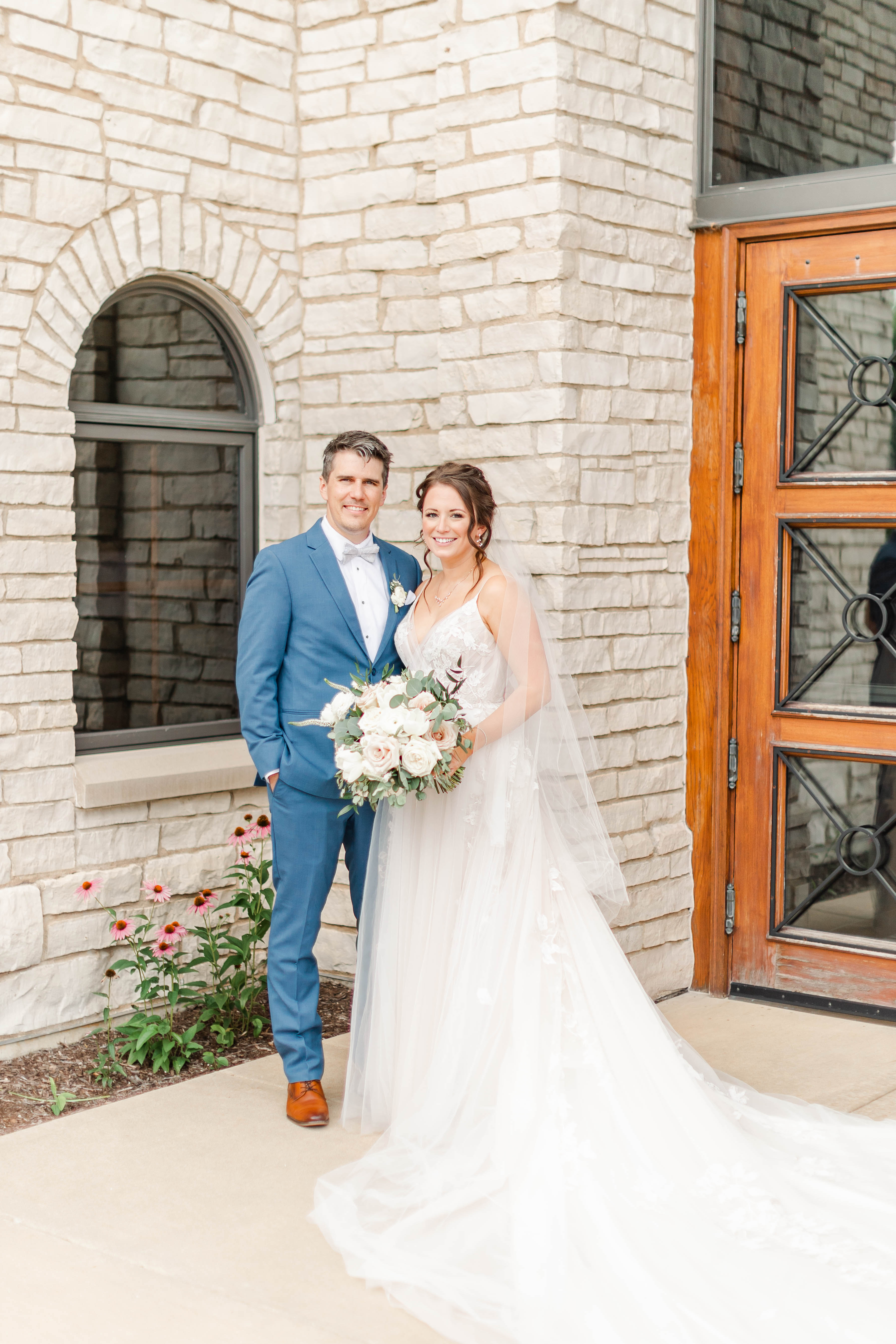 Groom in a medium blue suit and bride in a white wedding dress standing outside what appears to be a church.