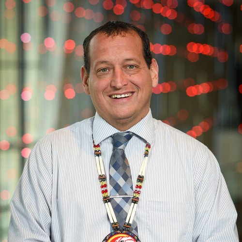 Mr Dropik wearing a light colored shirt and gray tie; with his ICS medallion. Colored red lights are in the background.