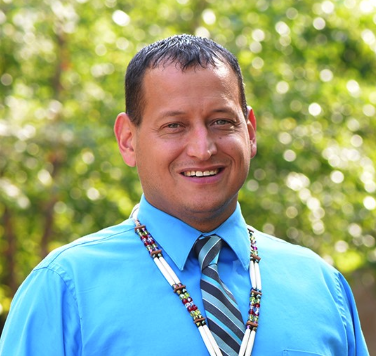 Mr Dropik smiling, wearing a blue collared shirt with a striped tie and beaded medallion. Green trees are in the background.