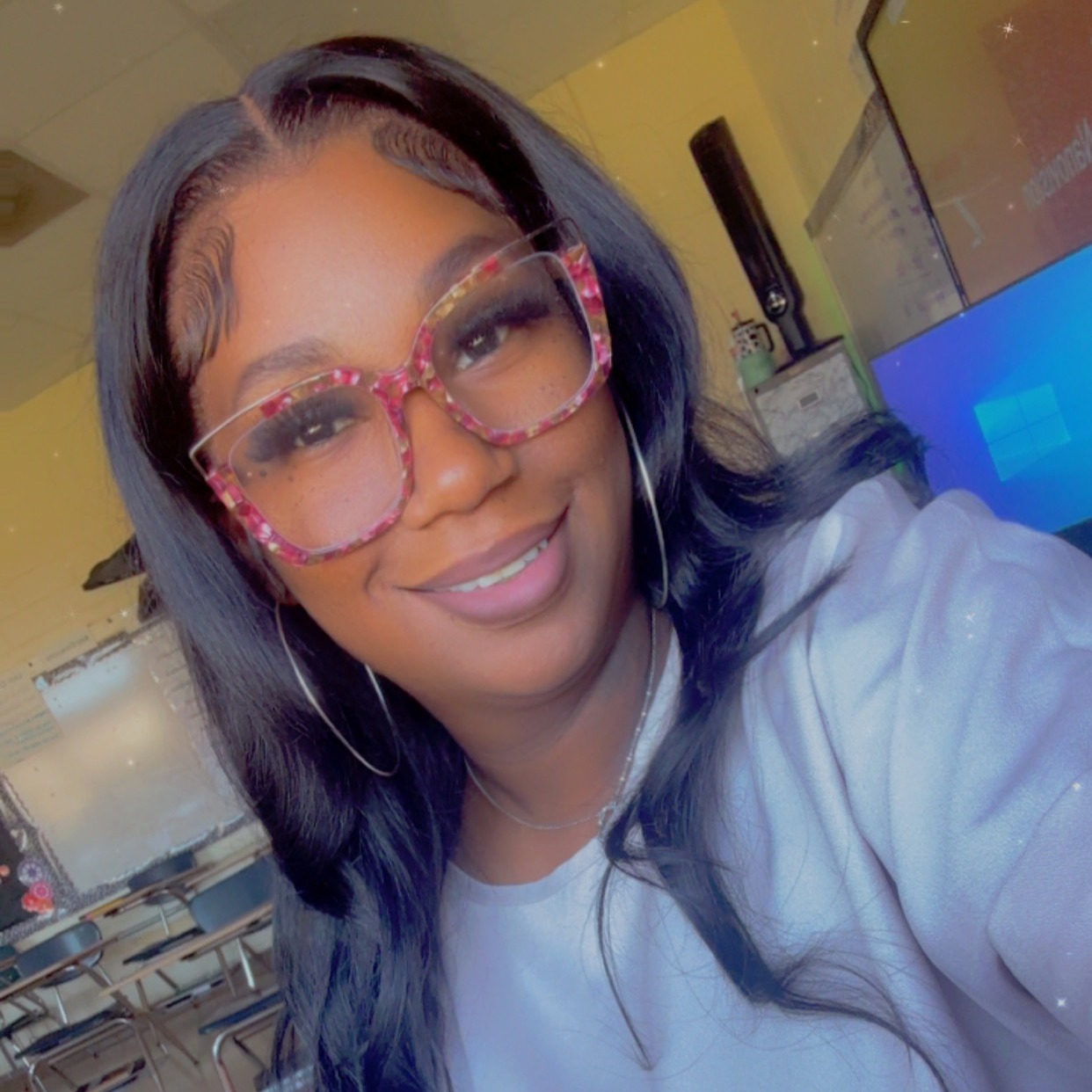 Woman with long hair wearing pink-framed glasses and a white shirt smiles in a classroom with yellow walls.